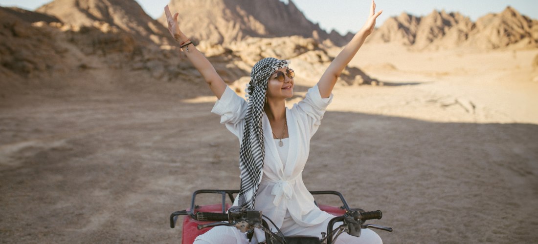 Eine lächelnde Frau sitzt auf einem roten Quad inmitten einer Wüstenlandschaft mit Felsen und Bergen im Hintergrund. Sie trägt weiße Kleidung, eine Sonnenbrille und ein schwarz-weißes Tuch um den Kopf. Die Arme hat sie weit nach oben gestreckt, als Ausdru