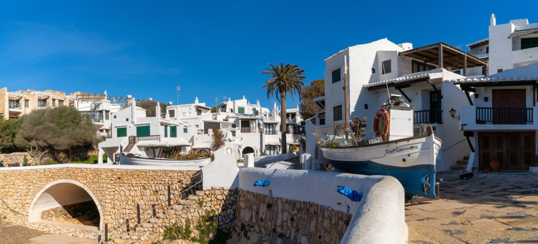 Panorama von Binibeca Vell mit weißen Häusern im traditionellen Fischerdorf-Stil, grünen Fensterläden, Palmen und einem Fischerboot im Vordergrund auf einer Steinrampe.