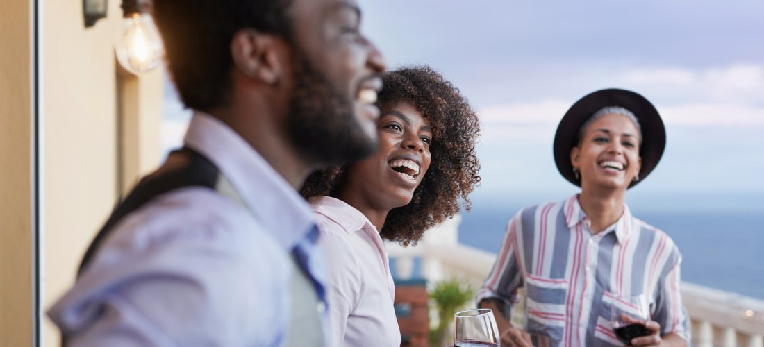 Drei Personen stehen auf einer Terrasse mit Meerblick, lachen und unterhalten sich. Zwei von ihnen halten Gläser mit Rotwein in der Hand. Die Stimmung ist gelöst und freundlich