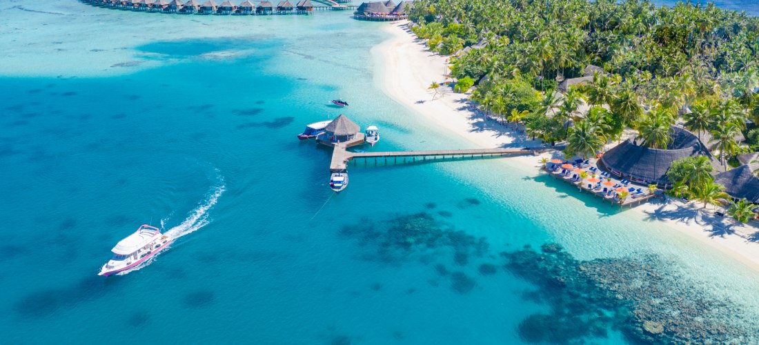 Luftaufnahme einer tropischen Insel auf den Malediven mit weißen Sandstränden, türkisblauem Wasser und palmengesäumten Ufer. Halbkreisförmig angeordnete Wasserbungalows ragen ins Meer. Mehrere Boote liegen an einem Steg, ein Schnellboot fährt durch das kl