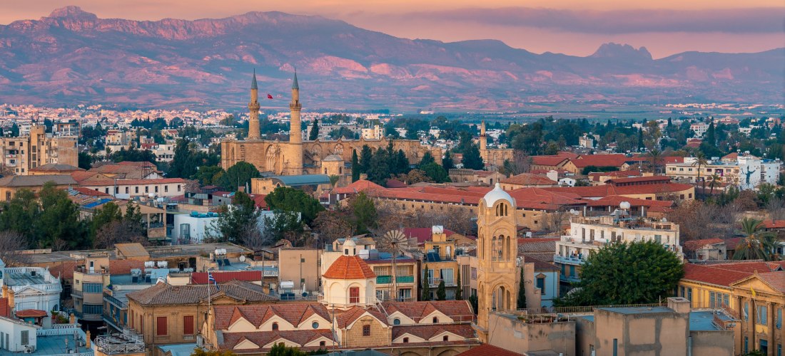 Panoramablick auf Nikosia (Lefkosia) bei Sonnenuntergang mit Stadt­dächern, Minaretten und dem Kyrenia-Gebirge im Hintergrund.