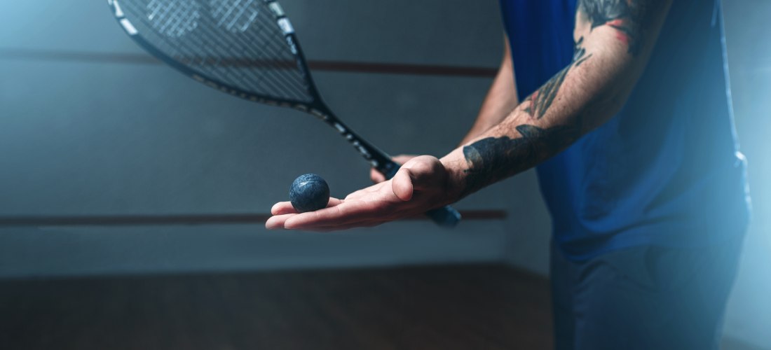 Nahaufnahme eines Squashspielers mit tätowiertem Arm, der einen Squashball in der geöffneten Hand hält und einen Schläger in der anderen. Der Hintergrund ist leicht verschwommen.