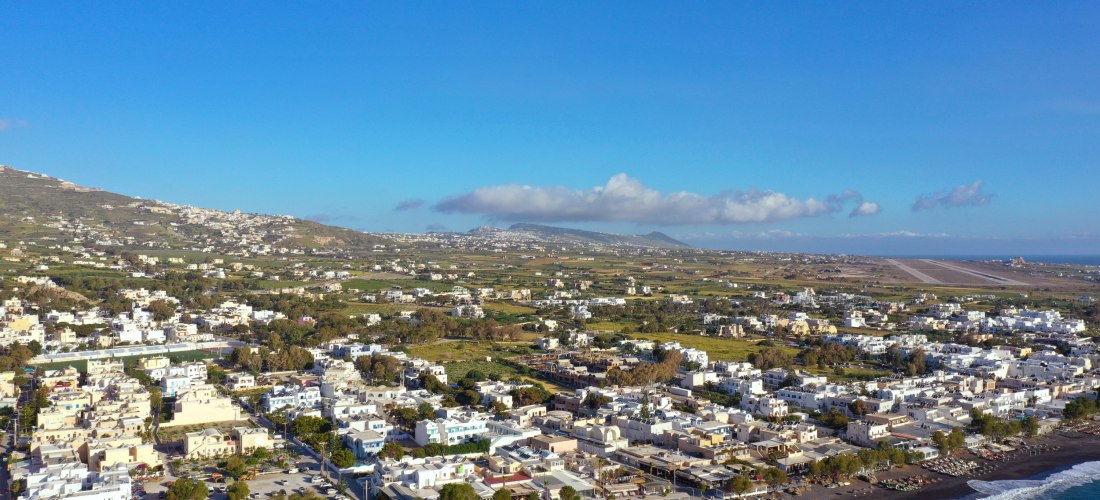 Luftaufnahme von Kamari auf Santorini: schwarze Strandlinie mit Liegen und Promenade, dahinter der Ort und die Start- und Landebahn des Flughafens