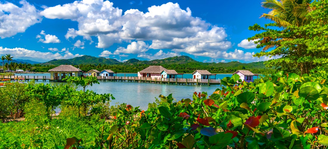 Blick auf charmante Bungalows auf Stelzen über dem Wasser, umgeben von grüner tropischer Vegetation mit Bergen im Hintergrund.