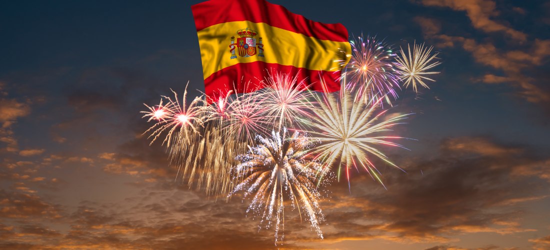 Spanische Flagge vor nächtlichem Himmel, umgeben von farbenfrohem Feuerwerk. Im Hintergrund geht die Sonne unter, der Himmel ist in orange und blau gefärbt
