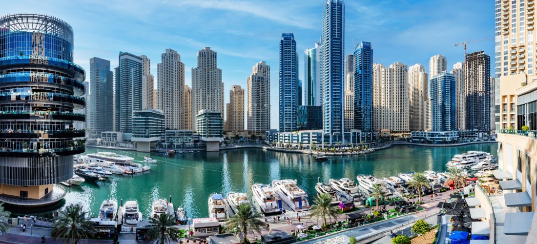 Panoramaaufnahme der Dubai Marina mit modernen Hochhäusern und zahlreichen Yachten im Wasser. Im Vordergrund verläuft eine elegante Uferpromenade mit Palmen, Cafés und Fußgängern. Links ist ein rundes, modernes Gebäude mit Glasfassade zu sehen. Der Himmel