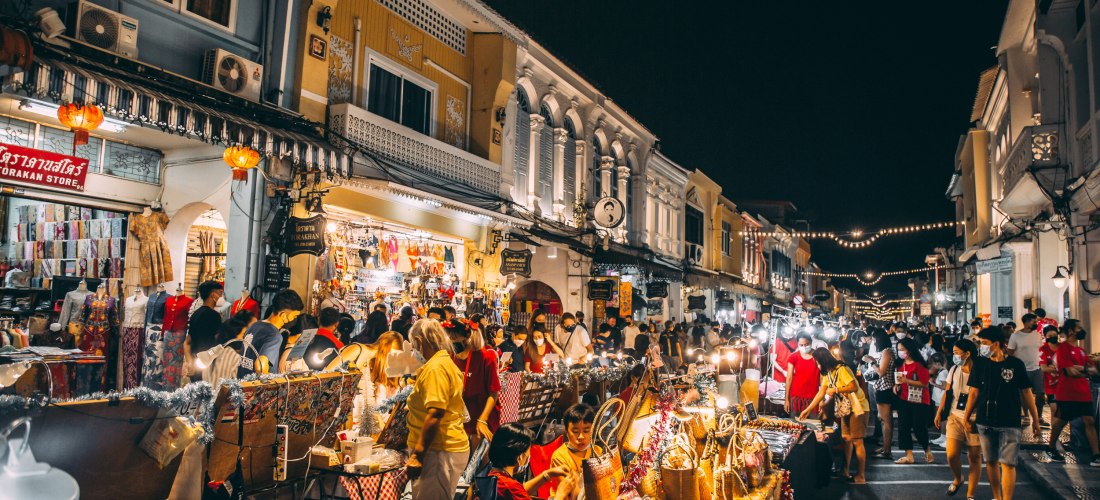 Lebendiger Nachtmarkt in Phuket Town bei Dunkelheit. Menschen schlendern durch die bunte Marktstraße, die von historischen Gebäuden mit Laternen und Lichterketten gesäumt wird. Zahlreiche Stände bieten Kunsthandwerk, Kleidung und Snacks an