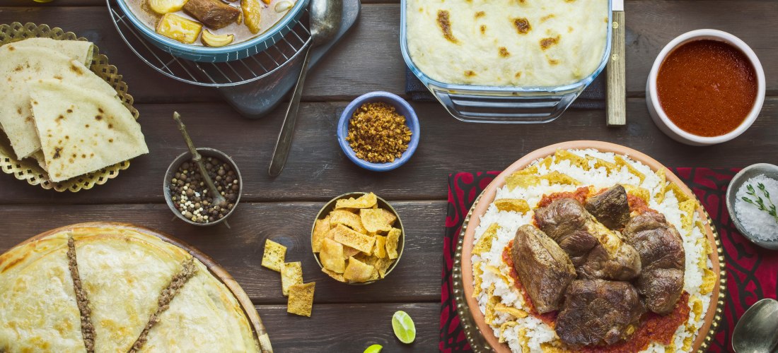 Orientalische Festtafel mit Lammfleisch auf Reis, Fladenbrot, Auflauf, Eintopf, Gemüse, Salat und Gewürzen auf einem Holztisch. Die Speisen sind vielfältig und farbenfroh angerichtet.