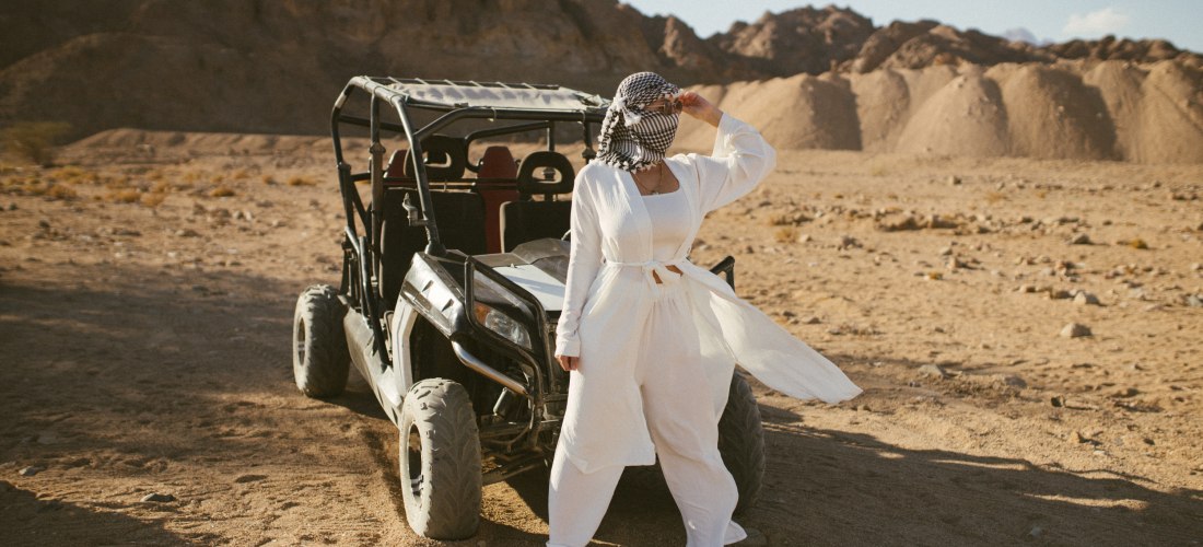 Eine Frau steht in einem weißen Outfit und mit Tuch um den Kopf neben einem Buggy in einer weiten, sonnigen Wüstenlandschaft. Im Hintergrund sind felsige Hügel zu sehen.