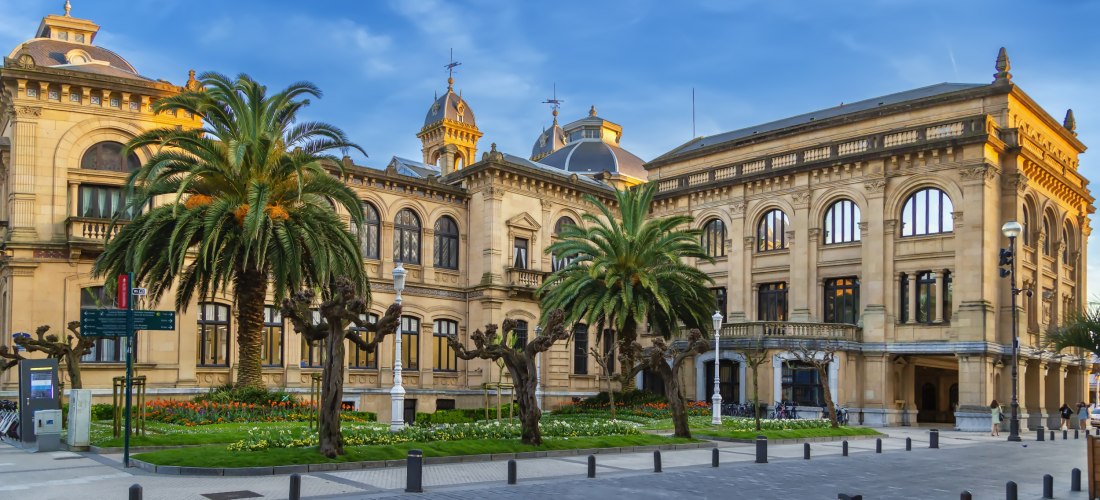 Das prächtige Rathaus von San Sebastián mit klassizistischer Architektur, Palmen und gepflegtem Blumenbeet im Vordergrund.