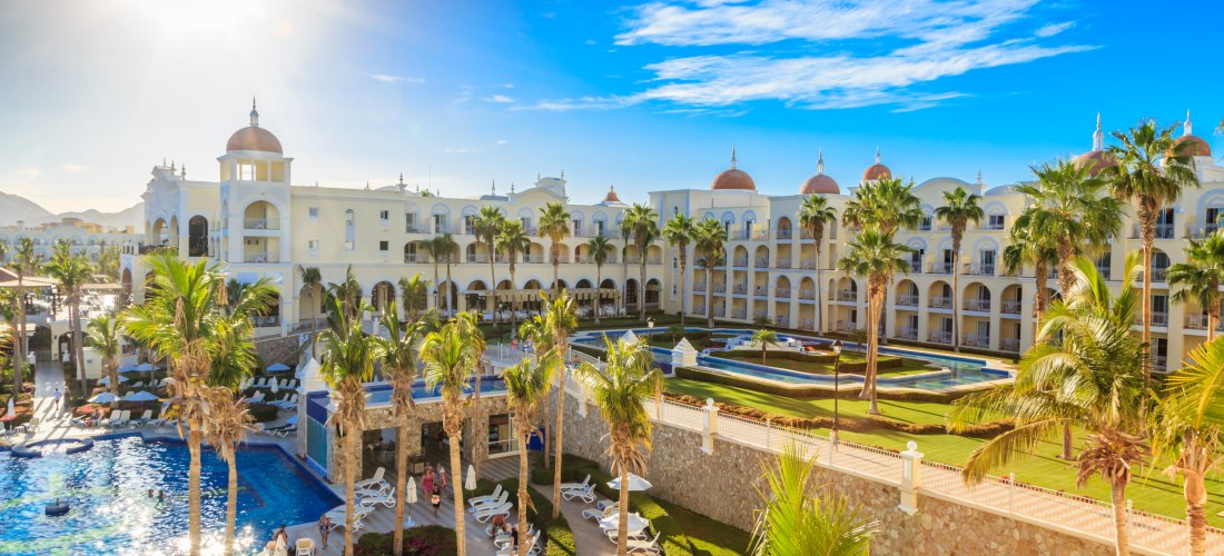 Sonnenbeschienenes, elegantes Hotelresort mit mehreren Etagen, weißer Fassade und Kuppeln. Im Vordergrund befinden sich Palmen, ein Poolbereich mit Liegestühlen sowie gepflegte Gartenanlagen. Der Himmel ist blau mit wenigen Wolken