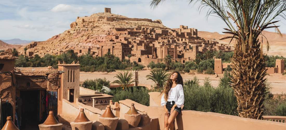 Frau lehnt entspannt an einer Mauer mit Blick auf die historische Lehmstadt Aït-Ben-Haddou in Marokko. Umgeben von Palmen, traditionellen Gebäuden und rötlich-gelben Felsen in der Wüstenlandschaft.