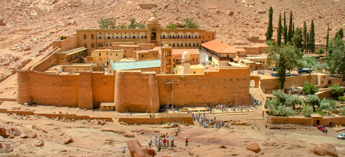 Das Katharinenkloster am Fuße des Berges Sinai, umgeben von rötlichen Felsen und karger Landschaft. Die massive Klosteranlage mit ihren hohen Mauern und Gebäuden in warmen Gelbtönen ist von zahlreichen Besuchern umgeben. Im Hintergrund ragen Zypressen und
