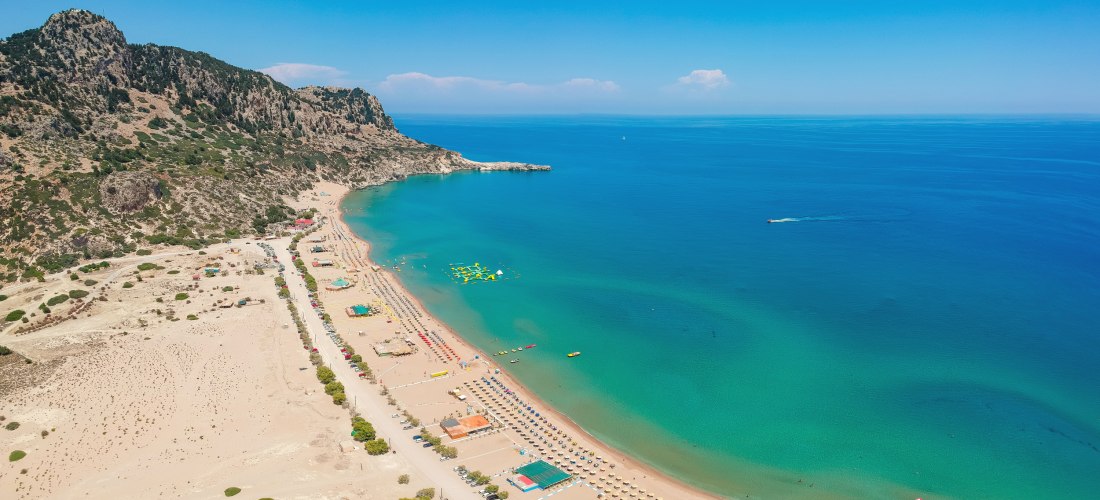 Luftaufnahme eines langen Sandstrandes mit klar türkisem Wasser an einem sanft geschwungenen Küstenabschnitt. Der Strand ist gut besucht, mit zahlreichen Sonnenschirmen und Liegen. Im Hintergrund ragt ein felsiges, grün bewachsenes Bergmassiv auf. Auf dem