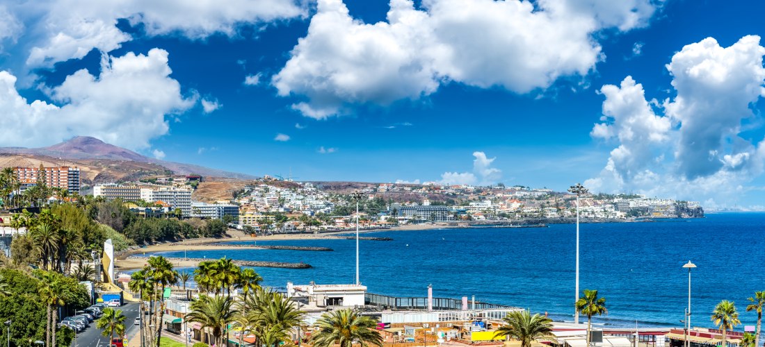 Blick auf San Agustín auf Gran Canaria mit Hotels, Apartmentanlagen und grünen Palmen entlang der Küste vor tiefblauem Atlantik und malerischem Wolkenhimmel.