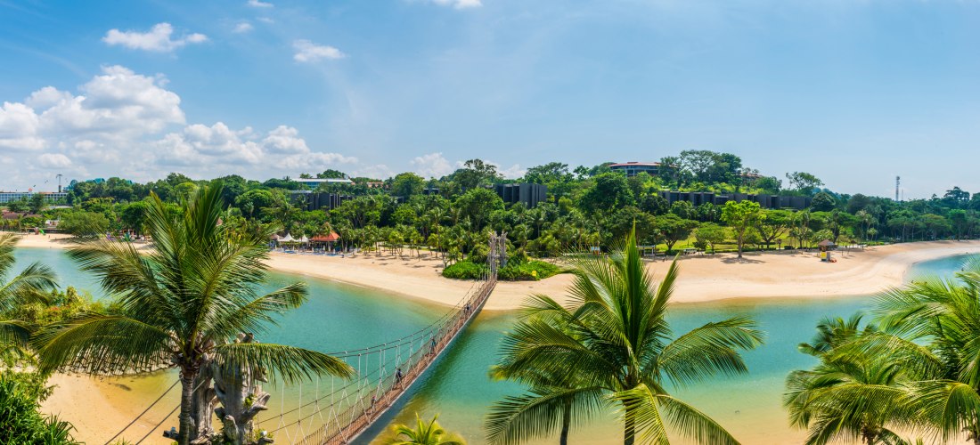 Panoramablick auf einen tropischen Strand auf Sentosa Island in Singapur. In der Mitte des Bildes verläuft eine schmale Hängebrücke über eine türkisfarbene Lagune, die von goldgelbem Sand und üppigen Palmen gesäumt ist. Im Hintergrund sieht man dichten, g