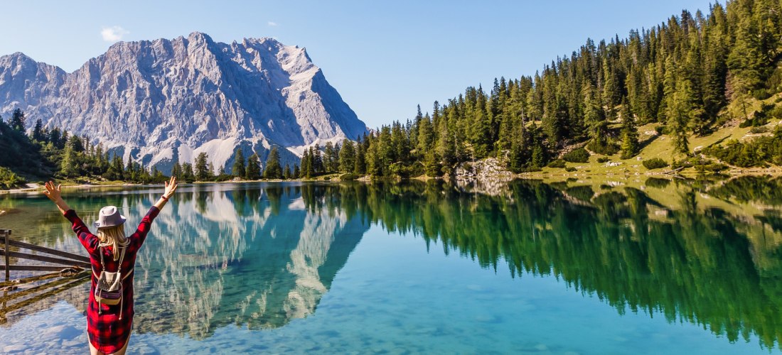 Alpensee mit spiegelglattem, türkisfarbenem Wasser; dahinter steile Felsberge und dichter Nadelwald. Im Vordergrund steht eine Person mit Rucksack und erhobenen Armen am Ufer.