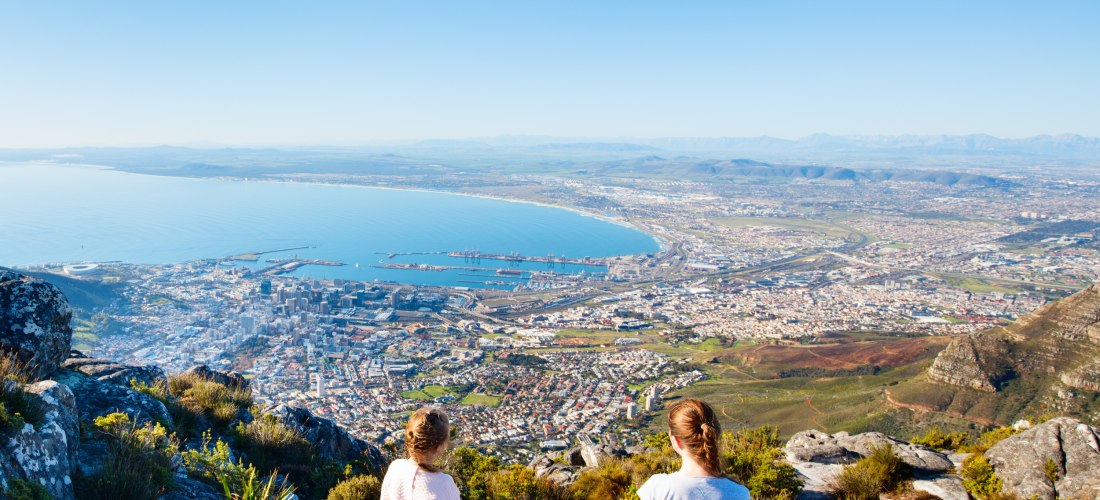 Zwei Personen sitzen auf einem Felsen hoch oben auf einem Berg und blicken auf die südafrikanische Stadt Kapstadt. Die Stadt liegt direkt am Meer, der Blick reicht über die Bucht bis zum Horizont. Es ist ein klarer, sonniger Tag, der Himmel ist wolkenlos 