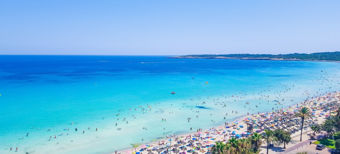 Breiter, heller Sandstrand mit vielen bunten Sonnenschirmen und Liegestühlen. Zahlreiche Menschen genießen die Sonne und das türkisfarbene Meer, das sanft in den Strand übergeht. Palmen und eine belebte Promenade mit Fußgängern, Autos und kleinen Geschäft