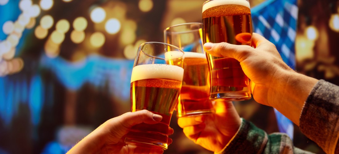 Drei Personen stoßen mit Biergläsern an. Im Hintergrund sind unscharfe, warme Lichter zu sehen, die eine festliche Stimmung erzeugen. Eine blaue Girlande ist leicht erkennbar