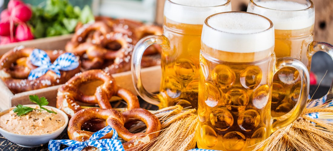 Drei Bierkrüge mit hellem Bier, daneben zahlreiche Brezeln, einige mit blau-weißen Schleifen, auf einem Tisch mit Weizenhalmen. Im Hintergrund sind Radieschen und frische Kräuter zu sehen – eine Szene im Stil des Oktoberfests