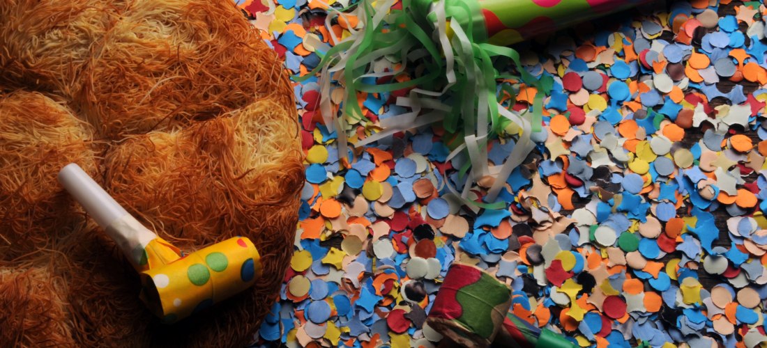 Buntes Konfetti bedeckt den Boden, daneben liegen Partytröten und ein großes, rundes Gebäck. Die Szene wirkt festlich und erinnert an Karneval oder eine Feier