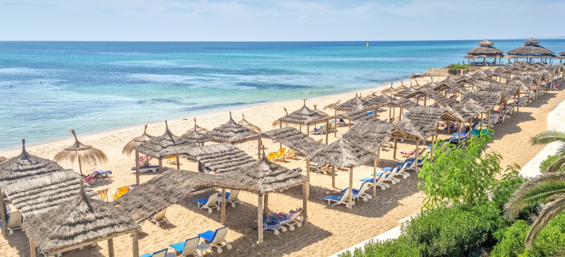 Ein langer, breiter Sandstrand mit feinem, goldgelbem Sand, gesäumt von einer Reihe Strohsonnenschirme und Liegestühlen. Im Hintergrund das türkisfarbene Meer, das sanft in verschiedenen Blautönen glitzert. Eine friedliche und einladende Strandatmosphäre.