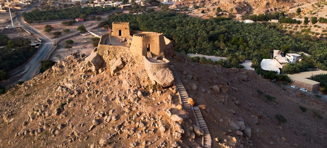 Luftaufnahme des Dhaya Fort in Ras Al Khaimah – historische Festung auf einem Hügel mit Palmenhainen und Gebirgspanorama.