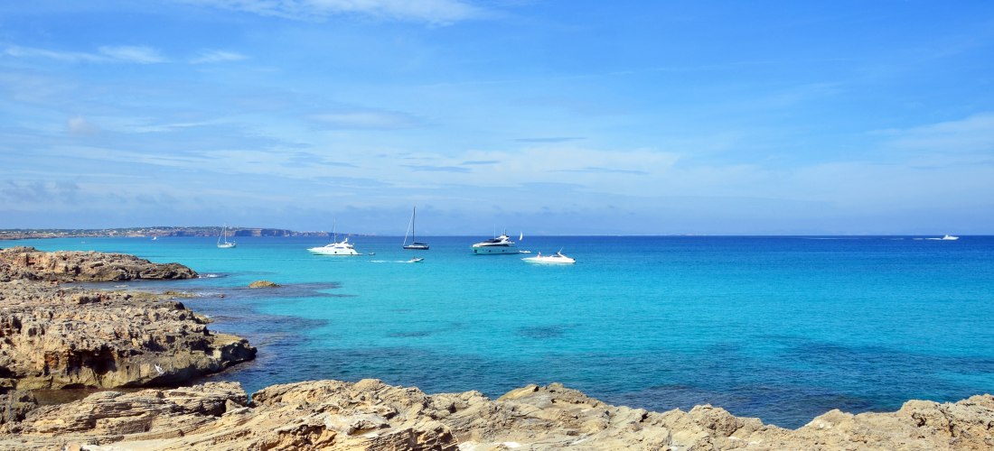 Felsige Küste auf Formentera mit weitem, türkisblauem Meer; mehrere Yachten und Segelboote liegen vor Anker, klarer Horizont unter leicht bewölktem, blauem Himmel.