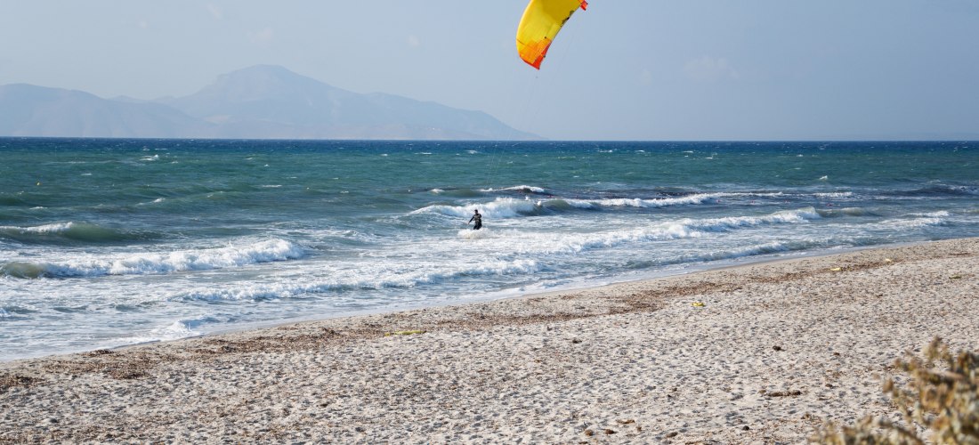 Mastichari Beach auf Kos: Kitesurfer mit gelb-rotem Schirm vor welligem, türkisblauem Meer und Bergkulisse.