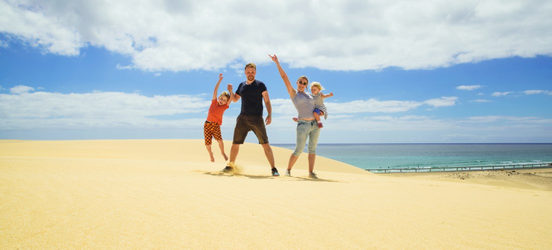 Lachende Familie mit zwei Kindern steht auf einer goldgelben Sanddüne vor blauem Himmel und Meer. Ein Kind wird hochgehoben, das andere auf dem Arm getragen.