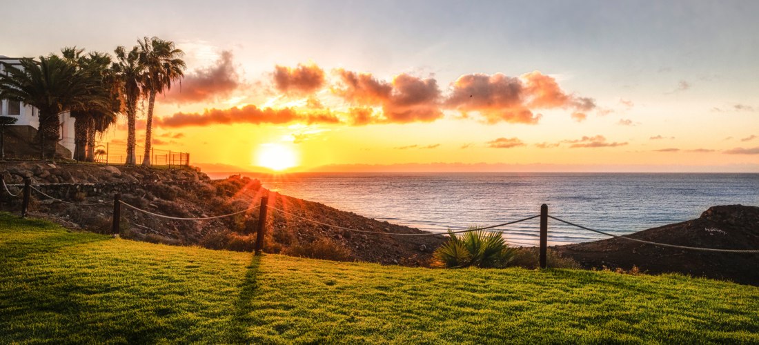 Sonnenuntergang über dem Meer, gesehen von einer grünen Rasenfläche mit Palmen und Felsen im Vordergrund. Der Himmel ist orange-rosa gefärbt mit vereinzelten Wolken.