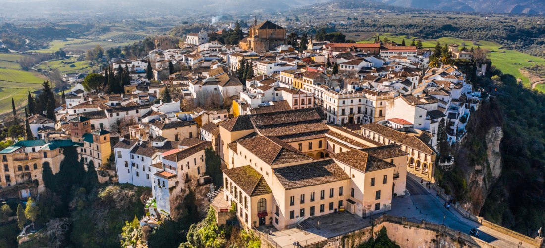 Luftaufnahme von Ronda in Andalusien, Spanien. Die weiße Altstadt liegt spektakulär auf einem Felsplateau, umgeben von grüner Landschaft und Bergen im Hintergrund.