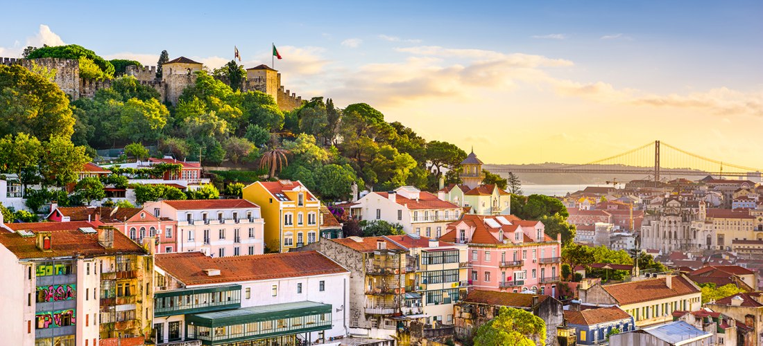 Panoramablick auf Lissabon bei Sonnenuntergang. Im Vordergrund farbenfrohe Häuser mit roten Dächern, im Hintergrund das Castelo de São Jorge auf einem bewaldeten Hügel. Rechts im Bild die Hängebrücke Ponte 25 de Abril über dem Tejo-Fluss.