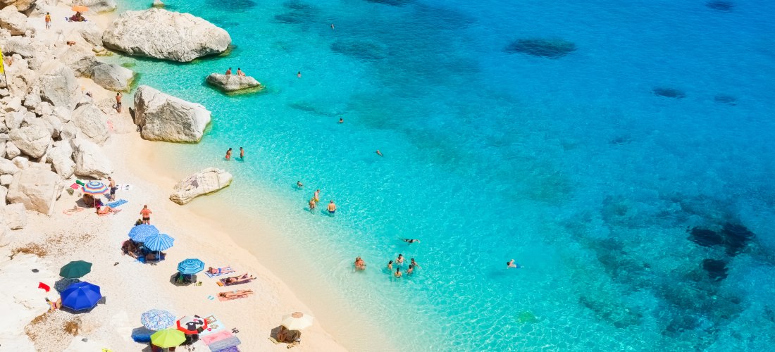 Menschen baden und entspannen an einem felsigen Strand mit türkisblauem Wasser und Sonnenschirmen auf hellem Sand