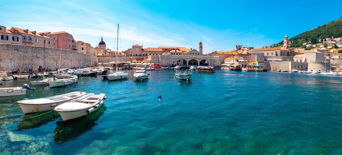 Kroatien - Dubrovnik Blick auf den alten Hafen von Dubrovnik mit kleinen Booten und Yachten im klaren, türkisfarbenen Wasser. Dahinter historische Gebäude mit roten Dächern und Stadtmauer, umrahmt von Hügeln.