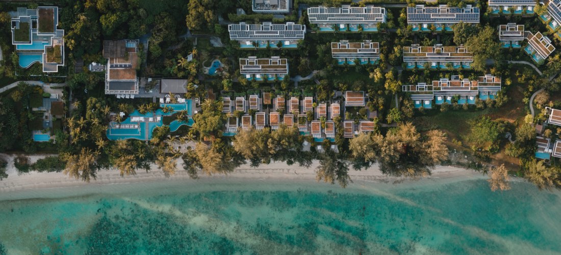 Luftaufnahme eines Resorts auf Phuket. Die Anlage liegt direkt am türkisfarbenen Meer, mit vielen Villen, Pools und tropischer Vegetation. Eine lange Strandlinie zieht sich entlang der gesamten Anlage.