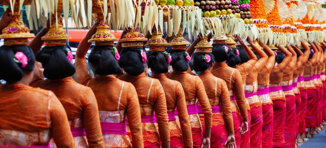 Eine lange Reihe balinesischer Frauen in traditioneller Kleidung mit orangefarbenen Blusen und pink-roten Sarongs tragen kunstvoll arrangierte Opfergaben auf ihren Köpfen. Die Gaben bestehen aus Früchten, Blumen und Palmenblättern. Jede Frau hat eine Blüt
