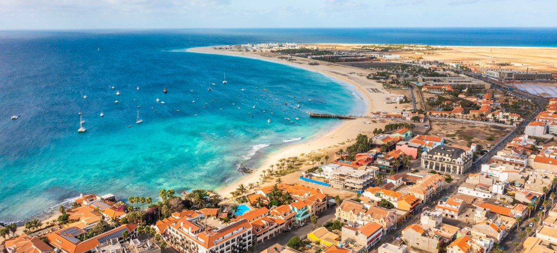 Luftaufnahme von Santa Maria auf Sal, Kapverden – türkisblaues Meer, halbmondförmiger Sandstrand, Boote in der Bucht und Stadt mit orangefarbenen Dächern