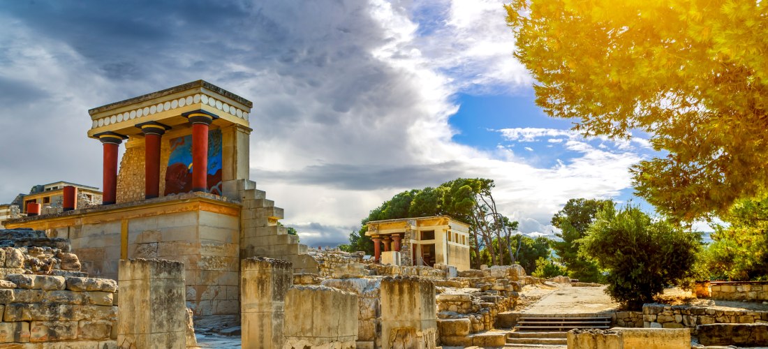 Blick auf die Ruinen des Palasts von Knossos auf Kreta. Im Vordergrund stehen antike Säulen und Steinmauern, in der Mitte ist ein restaurierter Gebäudeteil mit roten Säulen und farbigen Fresken zu sehen. Rechts scheint goldenes Sonnenlicht durch die Bäume