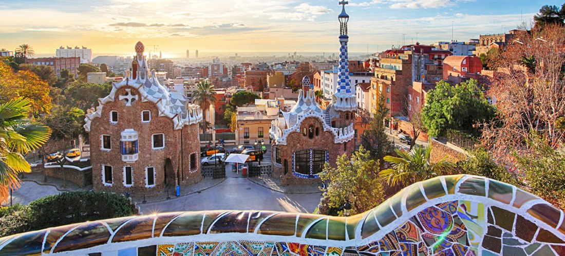 Blick über den Park Güell in Barcelona bei Sonnenaufgang. Im Vordergrund bunte Mosaikbänke, dahinter geschwungene Gebäude im Stil von Antoni Gaudí. Die Sonne geht über der Stadt auf, das Licht taucht den Park in warme Farben.