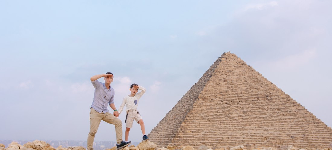Ein Mann und ein Junge stehen auf einem Steinhügel vor einer der Pyramiden von Gizeh in Ägypten. Beide blicken in die Ferne und halten sich mit einer Hand die Stirn gegen die Sonne. Die Szene wirkt wie ein gemeinsames Abenteuer oder Entdeckungsausflug.