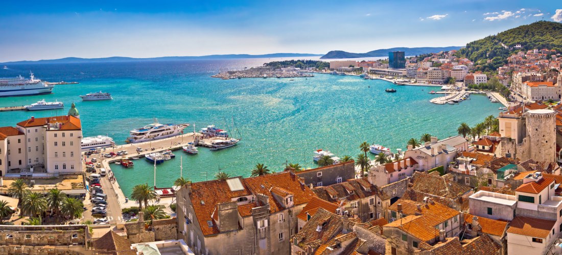 Panoramablick auf die Altstadt von Split in Kroatien mit roten Ziegeldächern, dem Hafen voller Yachten und Kreuzfahrtschiffen sowie türkisblauem Meer im Hintergrund. Entlang der Uferpromenade reihen sich Palmen und historische Gebäude.
