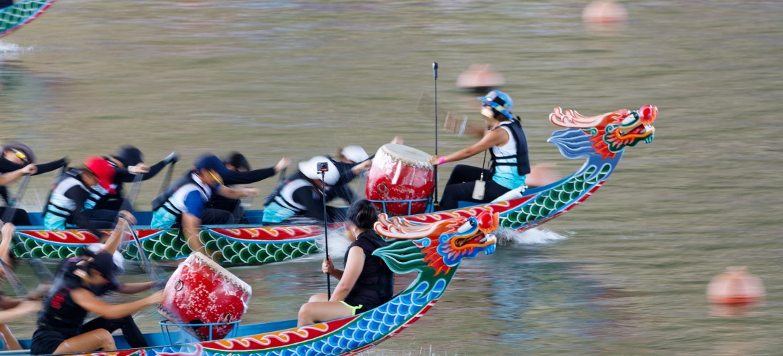 Drachenboote mit bunten, drachenförmigen Bugfiguren rasen im Wettkampf über das Wasser. Die Paddler bewegen sich synchron, während vorne je ein Trommler den Rhythmus vorgibt. Die Szene wirkt dynamisch und sportlich.