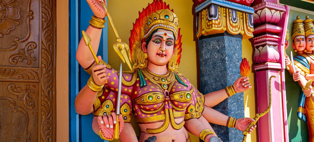 Dieses Bild zeigt eine beeindruckende, farbenfrohe Statue einer hinduistischen Göttin, vermutlich Durga oder Kali, die für Schutz und göttliche Macht steht. Die Figur hat mehrere Arme, von denen jeder ein Symbol oder eine Waffe hält, darunter ein Schwert,