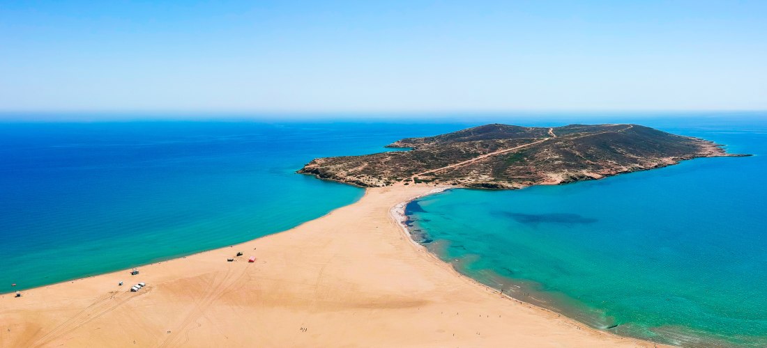 Luftaufnahme einer breiten Sandzunge, die zwei Meeresbuchten miteinander verbindet. Links und rechts leuchtet das Wasser in satten Türkis- und Blautönen. Die Halbinsel im Hintergrund ist karg und von rotbraunen Pfaden durchzogen. Der Strand ist weitläufig
