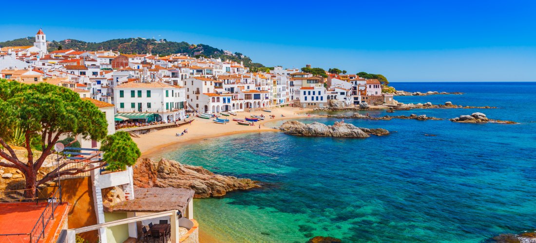 Blick auf ein malerisches Küstendorf an der Costa Brava mit weißen Häusern, Sandstrand und türkisfarbenem Meer. Einige Boote liegen am Ufer, dahinter bewaldete Hügel unter klarem, blauem Himmel.