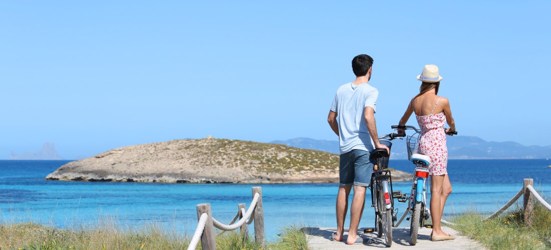 Ein Paar steht auf einem Holzsteg mit Fahrrädern und blickt auf das türkisblaue Meer und eine kleine Insel in der Ferne. Der Himmel ist klar und blau, und das Paar trägt sommerliche Kleidung