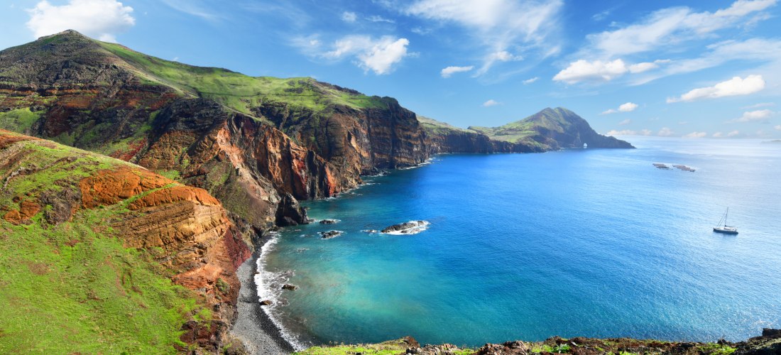 Steile, zerklüftete Küstenlinie mit rotbraunen Felsen und grünen Hängen auf Madeira, Portugal. Unten liegt eine kleine Bucht mit dunklem Kiesstrand, das Meer ist tiefblau und klar. Am Horizont ist ein Segelboot auf dem ruhigen Wasser zu sehen, darüber bla