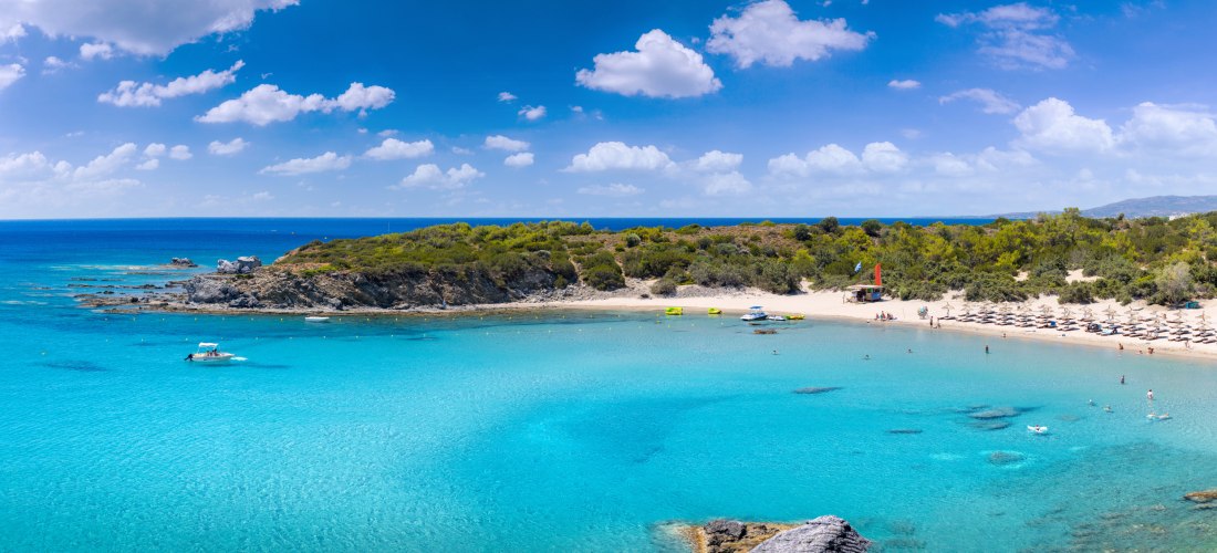 Das Bild zeigt eine paradiesische Lagune mit flachem, kristallklarem Wasser in hellen Türkis- und Blautönen.    Auf der rechten Seite verläuft ein weißer Sandstrand, bestückt mit zahlreichen Strohsonnenschirmen.    Einige Menschen baden oder entspannen im